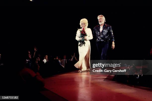 American Country musicians Dolly Parton, holding a bouquet of red roses, and Kenny Rogers walk onstage during a performance at Nassau Coliseum,...