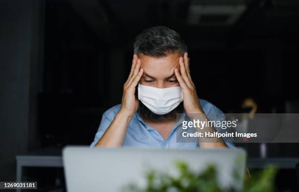 frustrated mature man with face mask sitting indoors in office, working. - emotional stress stock pictures, royalty-free photos & images