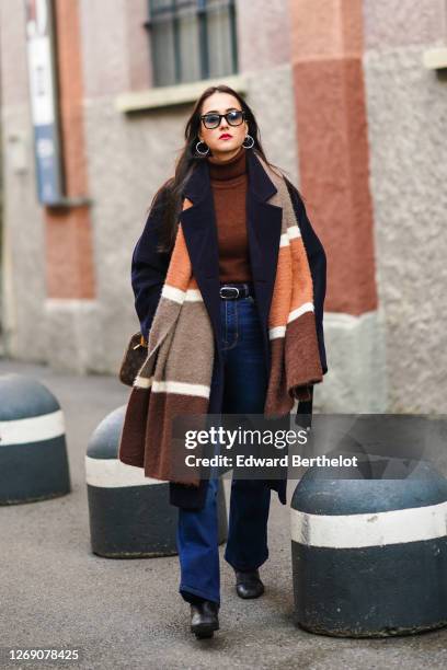 Guest wears sunglasses, earrings, a brown wool turtleneck pullover, a navy blue dark long coat, a brown and beige large scarf, a belt, blue denim...