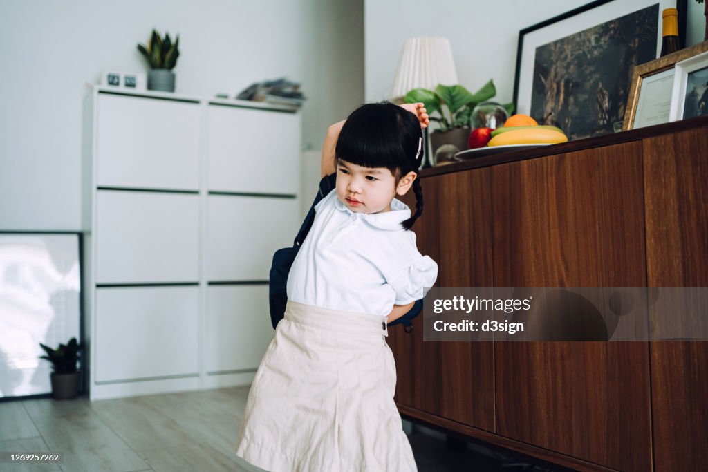 Little Asian girl with school uniform putting on school satchel at home and preparing for her first day at school