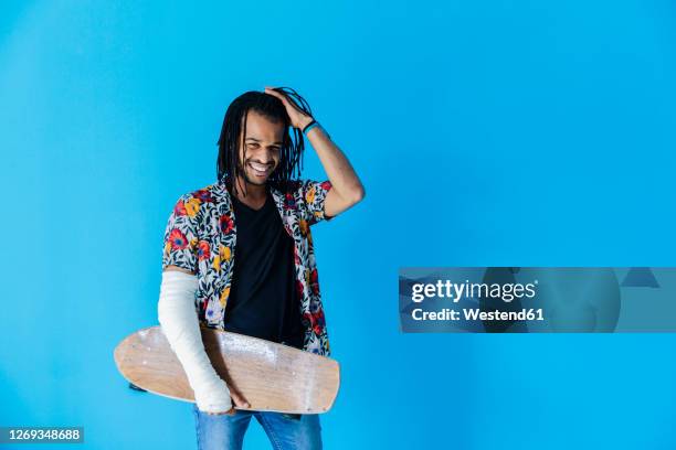 smiling man with fractured arm holding skateboard against blue background - broken skateboard stockfoto's en -beelden