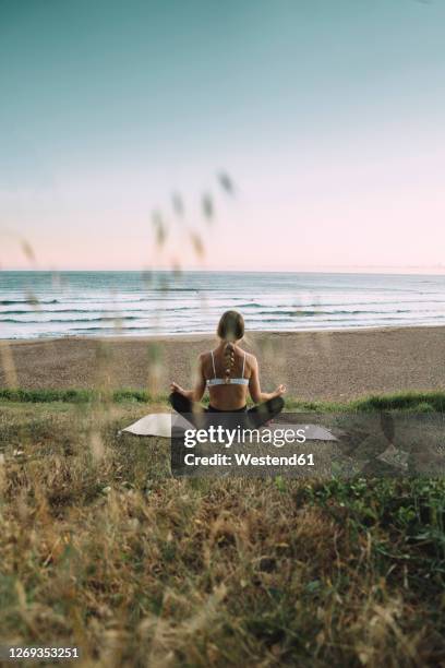 young woman meditating at beach against clear sky during sunset - strandmatte stock-fotos und bilder