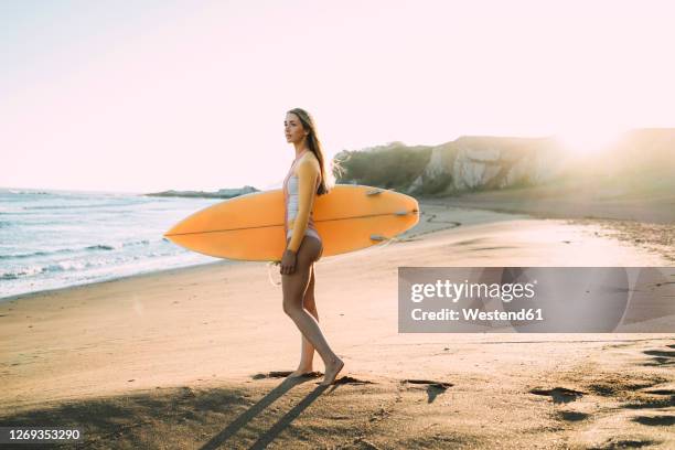 young woman wearing wetsuit carrying surfboard at beach during sunset - woman surfing stock pictures, royalty-free photos & images