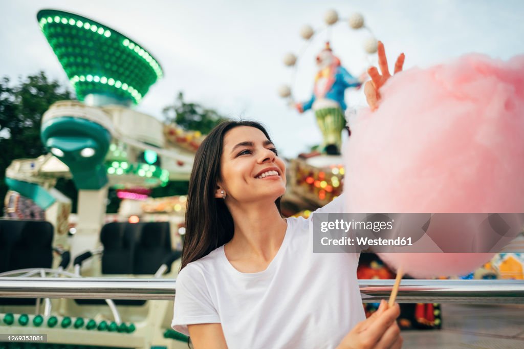 Smiling young woman holding cotton candy while standing at amusement park