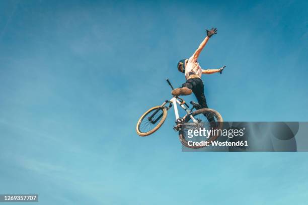 carefree man performing stunt with bicycle against blue sky during sunset - extreme sports bike stock pictures, royalty-free photos & images