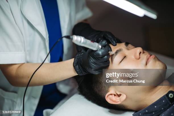 hommes chinois asiatiques beaux obtenant le traitement de sourcil de tatouage avec la machine au salon de beauté facial - maquillage permanent photos et images de collection
