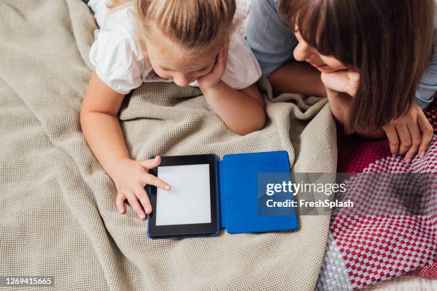 leer historias juntos: madre leyendo un libro en un e-reader a su hija en el dormitorio - lector de libros electrónicos fotografías e imágenes de stock