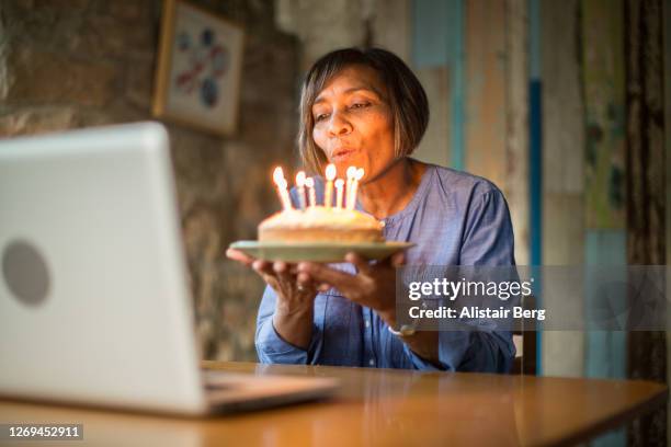 mature woman blowing out birthday cake candles on a video call - aufblasen stock-fotos und bilder