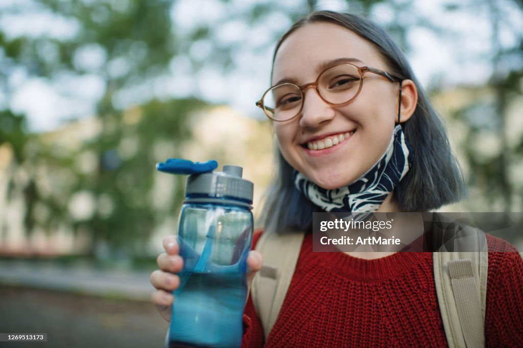 Teenager-Mädchen in wiederverwendbaren Schutz Gesichtsmaske Trinkwasser im Freien auf dem Campus oder Schulhof. Neues Normal- und Schulkonzept nach COVID-19-Quarantäne