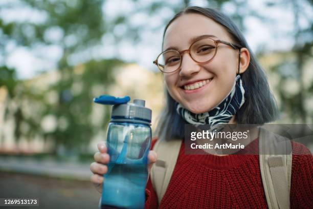 Teenage girl in reusable protective face mask drinking water outdoors in campus or schoolyard. New normal and Back to school concept after COVID-19 quarantine
