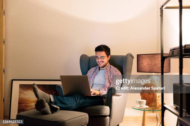 young man in glasses working at laptop at his apartment in the living room at night - comfortable chair stock pictures, royalty-free photos & images