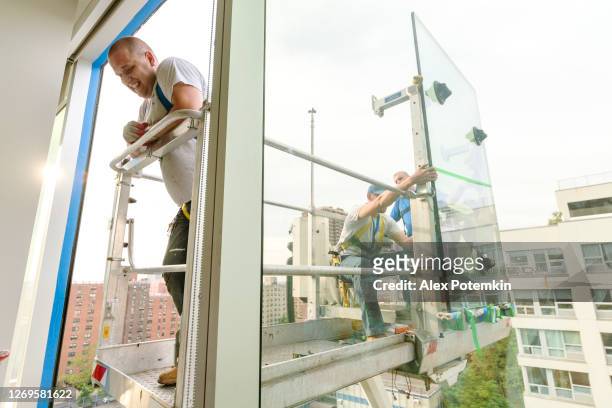 team of blue-collar workers replacing a broken window in the office building - applying a fresh sealant for a new glass installation. high-altitude work on the lifting platform which is placed outside. - replacement stock pictures, royalty-free photos & images