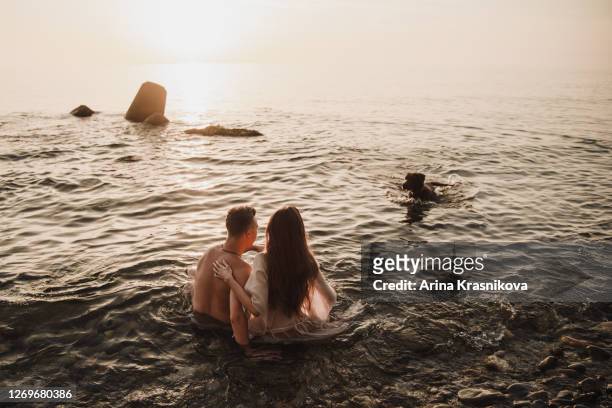 couple of young people sitting in the sea with a dog and watching the sunset - georgia love fotografías e imágenes de stock