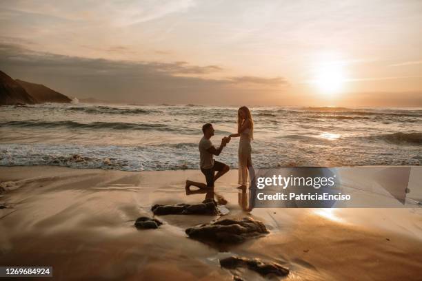 joven proponiendo a su novia en una playa - compromiso fotografías e imágenes de stock