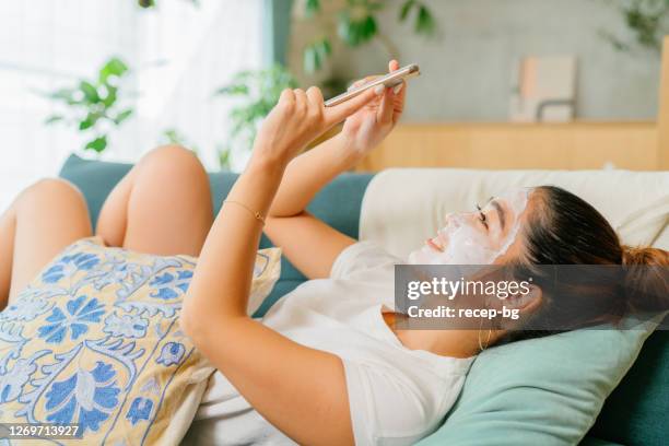 ソファに横たわり、自宅のリビングルームでスマートフォンを使って楽しむ顔の美しさのマスクを身に着けている女性 - フェイスパック ストックフォトと画像