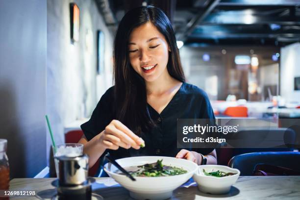 smiling young asian woman enjoying meal in a restaurant. she is squeezing fresh lime on her freshly served traditional vietnamese pho soup with noodles and beef, with vietnamese iced coffee by the side - vietnamese culture ストックフォトと画像