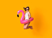 Happy young woman jumping on yellow background dressed in swimwear holding flamingo rubber ring beach.