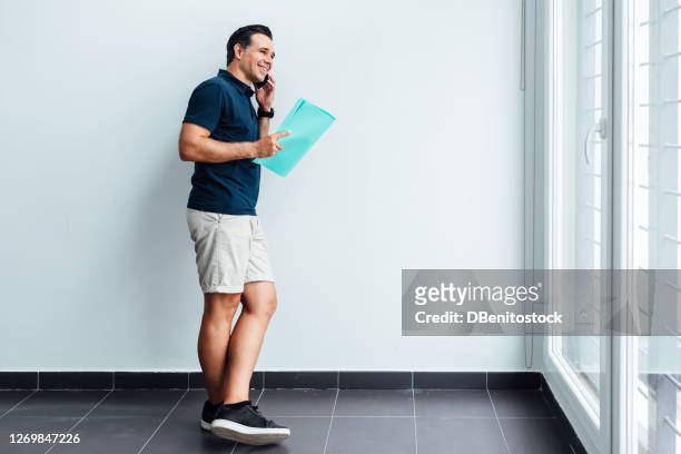 young entrepreneur man wearing a blue polo shirt, talking on the phone and smiling, holding an aquamarine folder next to a large window - ポロシャツ ストックフォトと画像