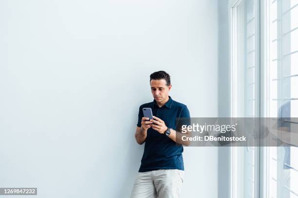 young entrepreneur man wearing a blue polo shirt looking at the mobile next to a large window - white polo shirt stock pictures, royalty-free photos & images