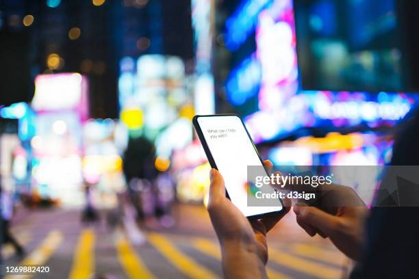 over the shoulder view of young woman using ai assistant on smartphone on the go in downtown city street, against city buildings with illuminated and multi-coloured neon commercial signs at night - ai assistant stock pictures, royalty-free photos & images