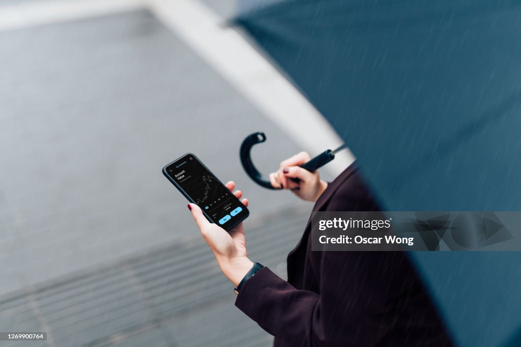 Businesswoman With Umbrella, Checking Stock Market On Smart Phone In The Rain