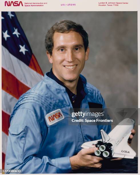 American NASA astronaut Michael J Smith , wearing a blue NASA jumpsuit, smiling in a studio portrait while holding a model of a space shuttle at...
