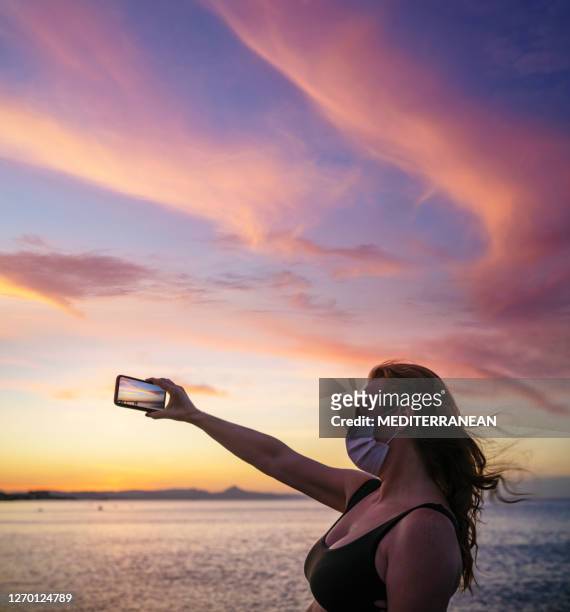 turista adulto de los años 50 mujer en pandemia máscara de la cara en la playa del atardecer tomando fotos - vista marina fotografías e imágenes de stock