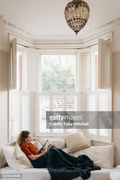 young woman relaxing on living room sofa reading book in bay window - fensterladen stock-fotos und bilder