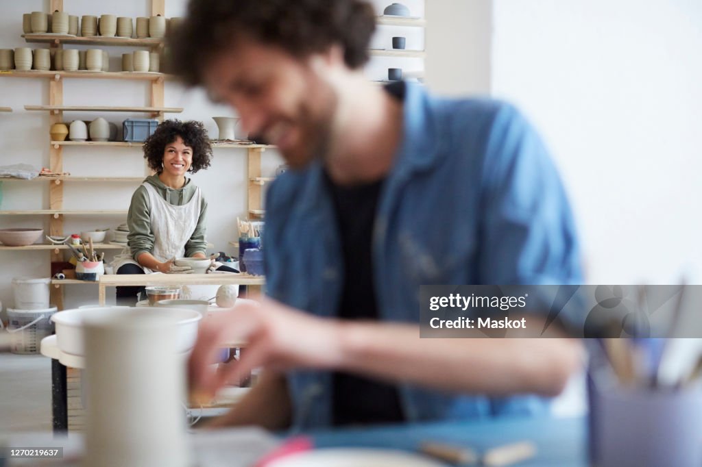 Portrait of smiling woman learning pottery while man in foreground at art studio