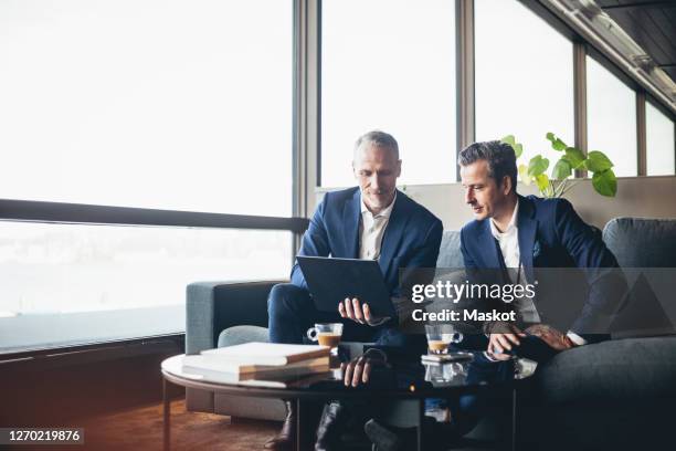 businessman showing laptop to colleague while sitting on sofa at workplace - completo da uomo foto e immagini stock