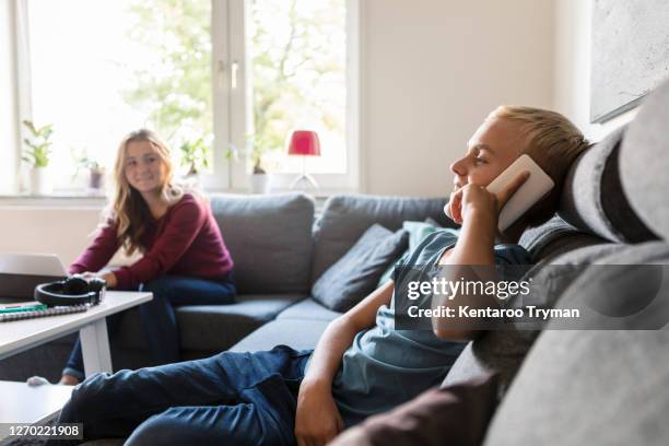 male teenager talking on phone while smiling sister sitting on sofa at home - male teen tablet stock-fotos und bilder