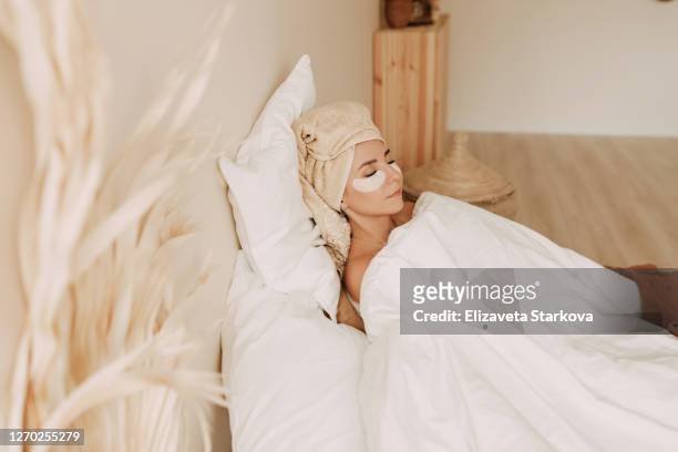 an adult woman with a towel on her head and patches on her face relaxes in bed under a blanket - one eyed stock pictures, royalty-free photos & images