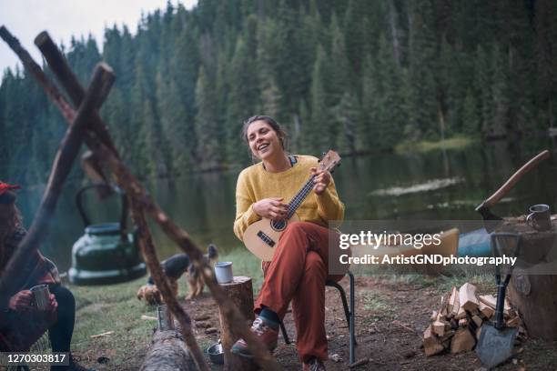 jeune femme gaie s’amuse sur ses vacances de camping tout en jouant ukulele à côté du feu de camp - sing outside photos et images de collection