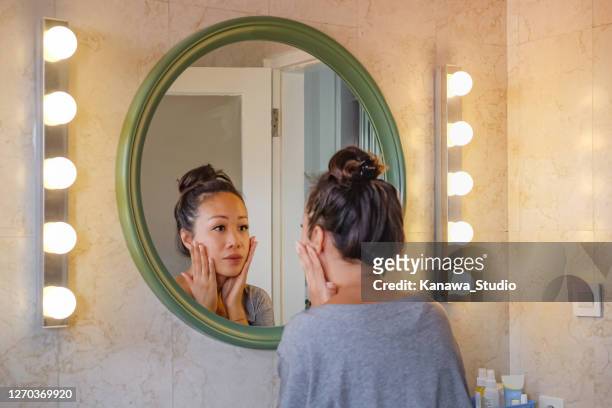worried east asian woman checking her face skin in the mirror. - looking stock pictures, royalty-free photos & images