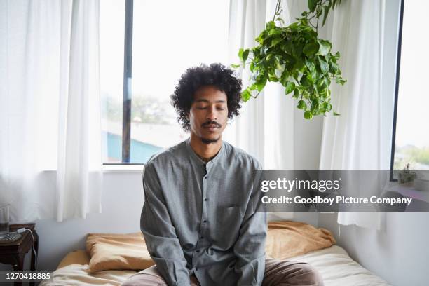 young man sitting on his bed and meditating with his eyes closed - buddhism prayer stock pictures, royalty-free photos & images