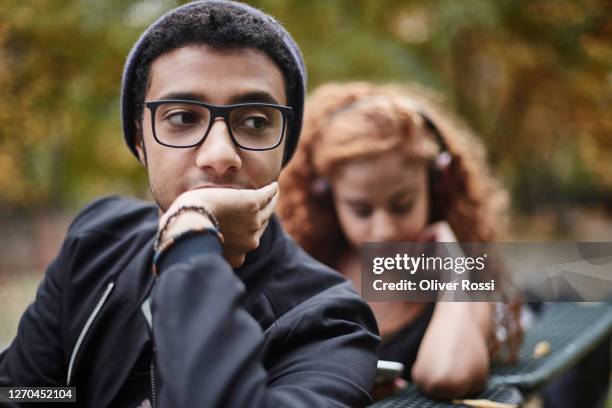serious young man on park bench with girlfriend in background - ignorance photos et images de collection