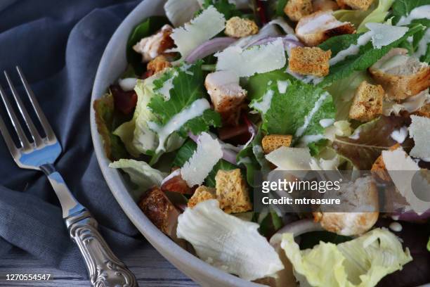close-up image of chicken caesar salad in bowl, lettuce, red onion, beetroot leaves, cubed bacon, roasted chicken, topped with croutons and parmesan cheese shavings, besides grey tea towel and metal fork, wood grain background, elevated view - shaved parmesan cheese stock pictures, royalty-free photos & images