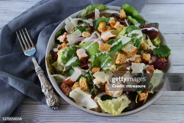image of chicken caesar salad in bowl, lettuce, red onion, beetroot leaves, cubed bacon, roasted chicken, topped with croutons and parmesan cheese shavings, besides grey tea towel and metal fork, wood grain background, elevated view - caesar salad stock pictures, royalty-free photos & images