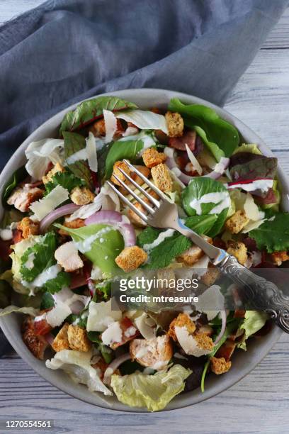 image of chicken caesar salad in bowl, lettuce, red onion, beetroot leaves, cubed bacon, roasted chicken, topped with croutons and parmesan cheese shavings, with metal fork besides grey tea towel, wood grain background, elevated view - shaved cheese stock pictures, royalty-free photos & images