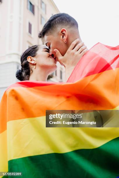 portrait of young lesbian couple standing on a street, wrapped in rainbow flag, kissing. - photos of lesbians kissing stock pictures, royalty-free photos & images