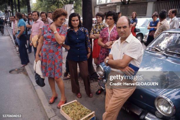 Groupe de femmes près d'un homme adossé à une voiture vendant du raisin dans une cagette posée à terre, dans un parking à l'entrée de l'usine Fiat,...