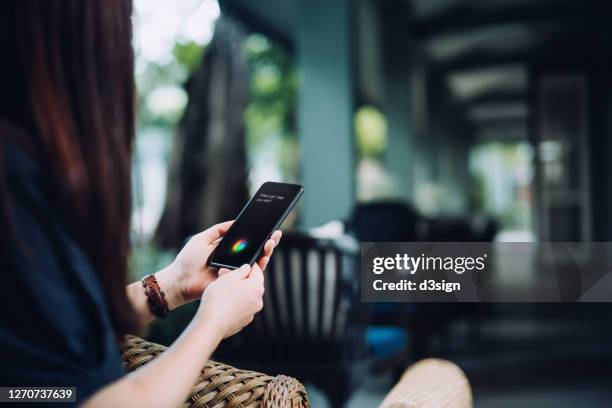 over the shoulder view of young asian female traveller using ai assistant on smartphone while on staycation relaxing in a hotel - speech recognition stockfoto's en -beelden