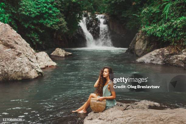 young pretty woman with long black curly hair sitting with view of natural water lagoon of waterfall in bali. handmade wooden handbag and summer green dress. stylish and trendy look. - women in see through dresses stock-fotos und bilder