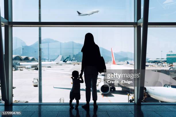 silhouette of joyful young asian mother holding hands with cute little daughter looking at airplane through window at the airport while waiting for departure - emigration and immigration foto e immagini stock