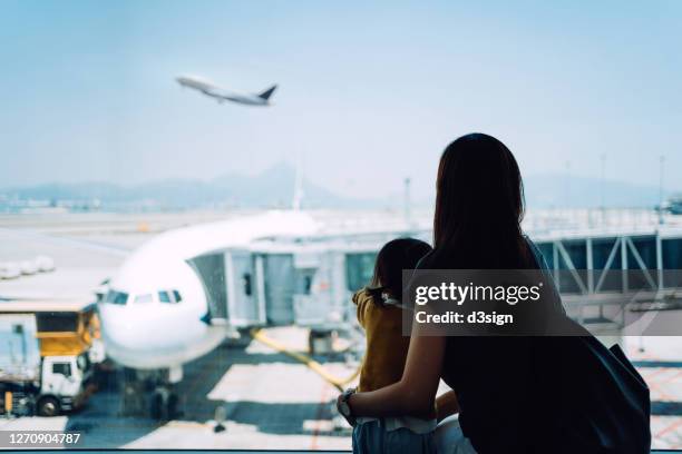 young asian mother embracing her cute little daughter looking at airplane through window at the airport while waiting for departure - baby arrival stock pictures, royalty-free photos & images