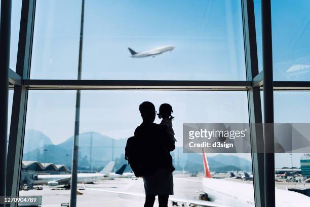 silhouette of joyful young asian father carrying cute little daughter looking at airplane through window at the airport while waiting for departure - emigration and immigration foto e immagini stock