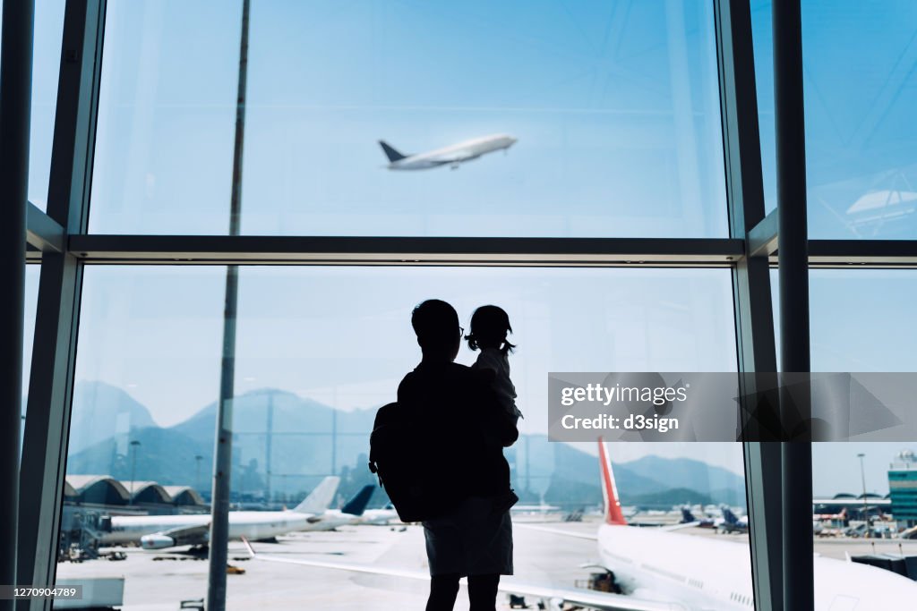 Silhouette of joyful young Asian father carrying cute little daughter looking at airplane through window at the airport while waiting for departure