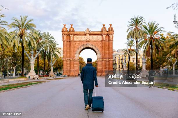 rear view of a man with suitcase walking towards arc de triomf in barcelona, spain - winter barcelona stock pictures, royalty-free photos & images