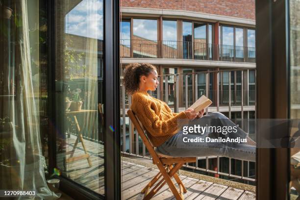 junge frau liest ein buch zu hause - balcony stock-fotos und bilder