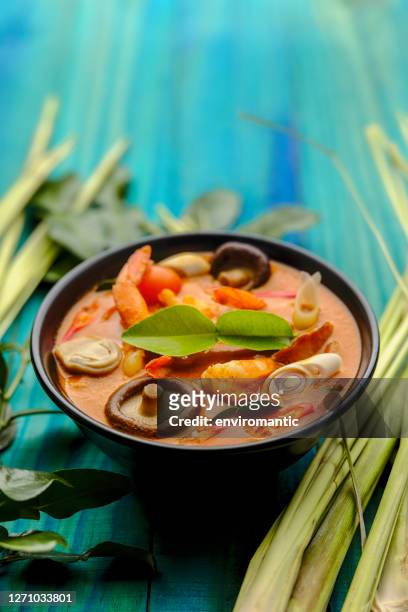 berühmtes, international bekanntes würziges, kokosnuss-thai-suppengericht von 'tom yam kung', in einer schüssel neben ganzem zitronengras und kaffir-limonenblättern auf einem türkisfarbenen holztisch-hintergrund. - curry soup stock-fotos und bilder